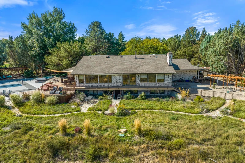 A large stone house with a wraparound deck overlooking a green field and trees under a blue sky.