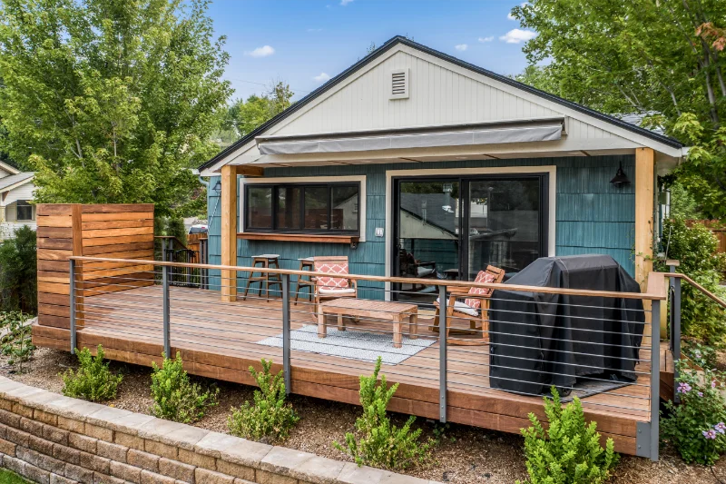 Wooden deck with a blue-sided house, awning, and grilling setup; low brick retaining wall with landscaping.