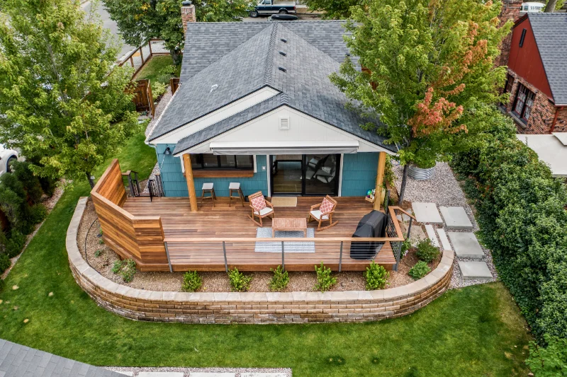 Aerial view of a blue house with a wooden deck, retaining wall, and green lawn.