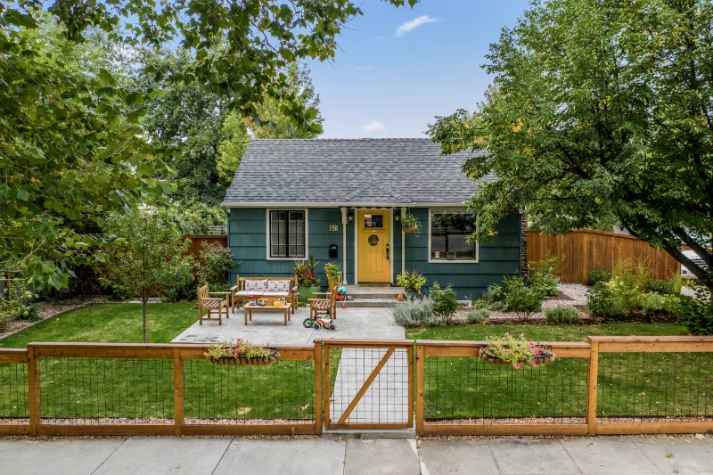 Teal cottage with yellow door, small front patio, and wooden fence.