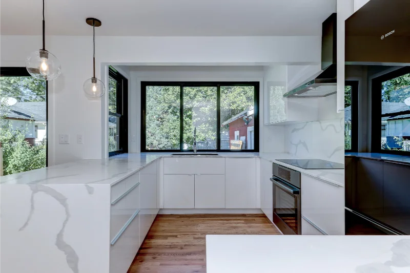 Modern white kitchen with marble countertops, windows, and stainless steel appliances.