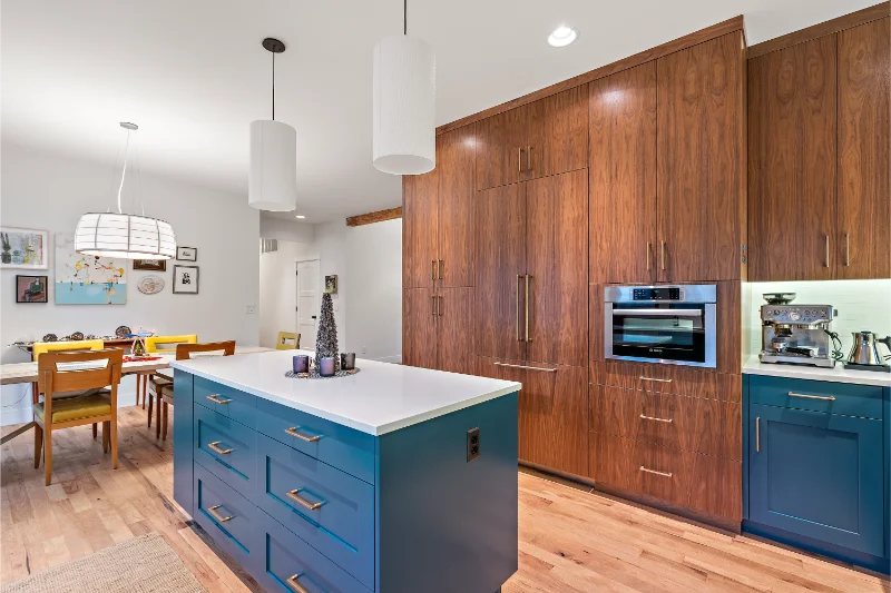 Modern kitchen with blue island, wood cabinets, oven, and dining area with yellow chairs.