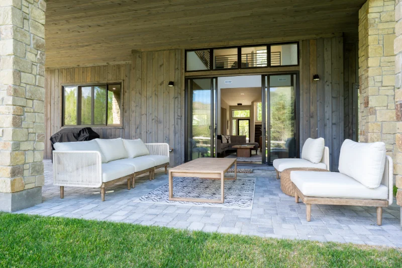 Patio with light-colored sofa and chairs, wooden table, and sliding glass door. Overhang with stone columns, and green grass.