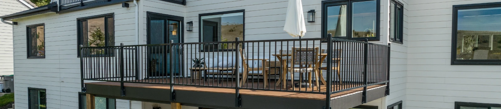 A modern house exterior with a deck, black railings, and white brick walls. The deck has a small table, chairs, and umbrella.