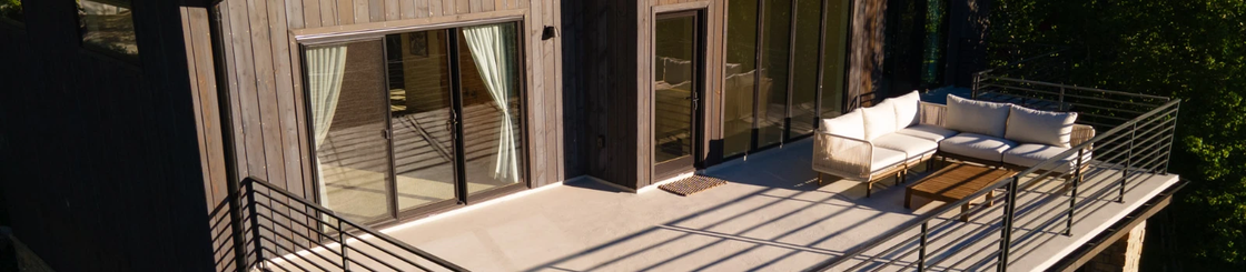 Wooden cabin with outdoor seating area. Cream couch, coffee table, and metal railing on the deck. Sliding glass doors.