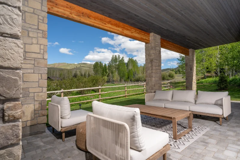 Patio with light-colored sofas, wooden table, and scenic view of green fields and trees under a cloudy sky.