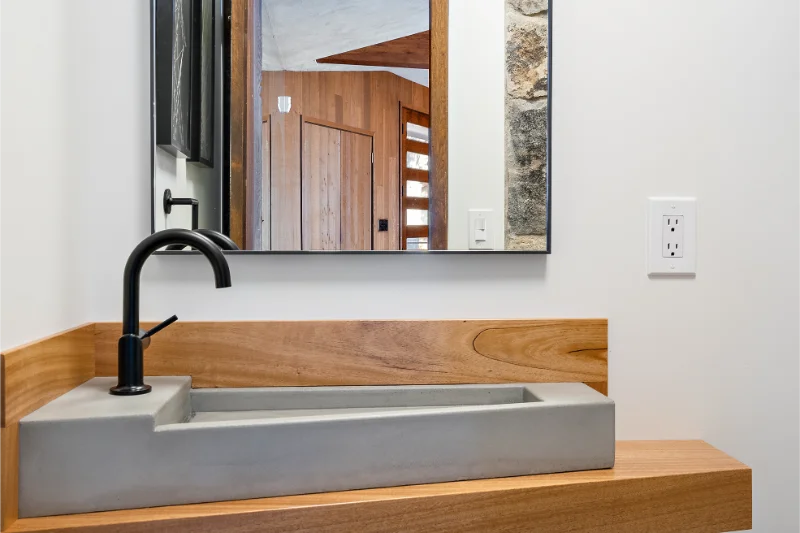 Modern bathroom with gray concrete sink, black faucet, and wood accents. Mirror reflects wooden door.