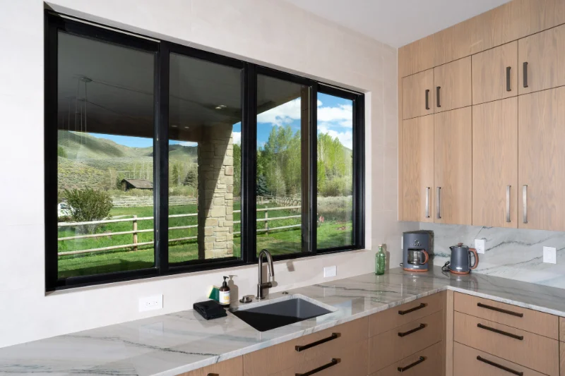 Kitchen with large window overlooking a green landscape and wood cabinets with a light countertop.