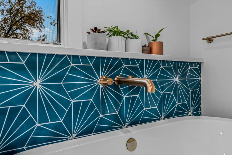 Blue geometric tile backsplash above a white bathtub with gold faucet, window, and plants.