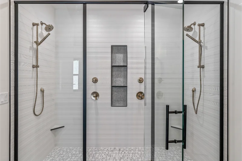 Double shower with glass doors, white tiled walls, pebble floor, and dark metal fixtures.