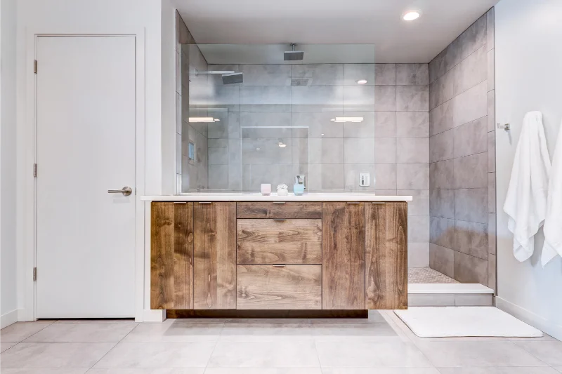 Bathroom with wooden vanity, glass shower, and gray tile walls. White door and towels on the right.