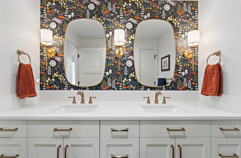Bathroom with floral wallpaper, two oval mirrors, gold fixtures, white vanity, and rust-colored towels.