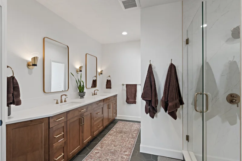 Bathroom with double vanity, gold mirrors, brown cabinets, and a glass shower.