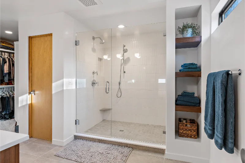 Modern bathroom with a glass shower, wooden shelves holding towels and plant, and a walk-in closet.
