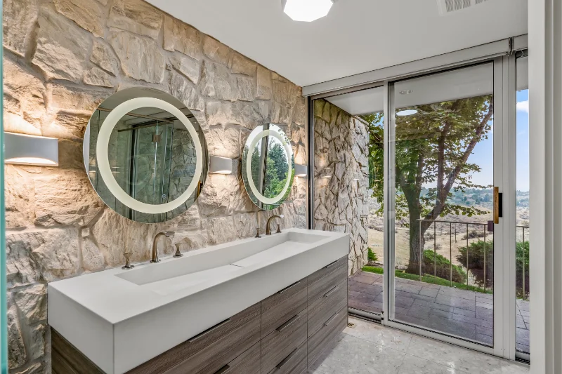 Bathroom with stone walls, double vanity, round mirrors, and glass sliding door overlooking trees.