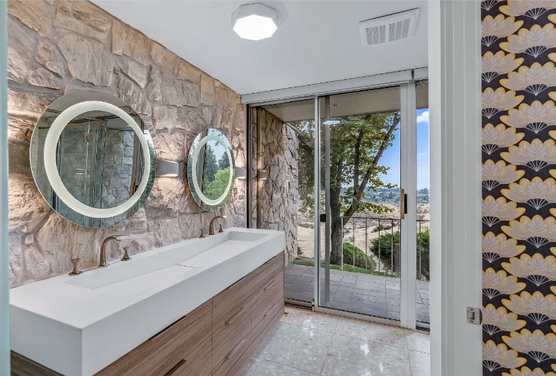 Modern bathroom with stone wall, rectangular sink, round mirrors, wood cabinet, sliding glass door with a view.