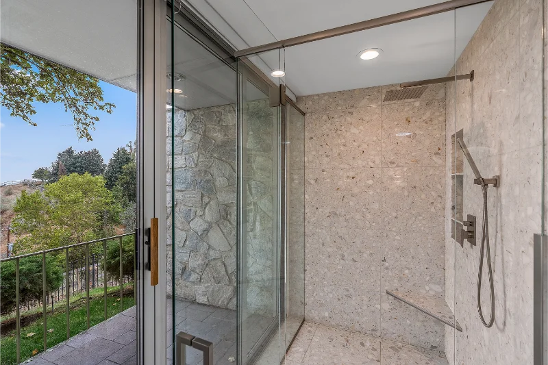 Modern bathroom with glass shower, stone walls, and sliding glass door to a balcony with green trees.