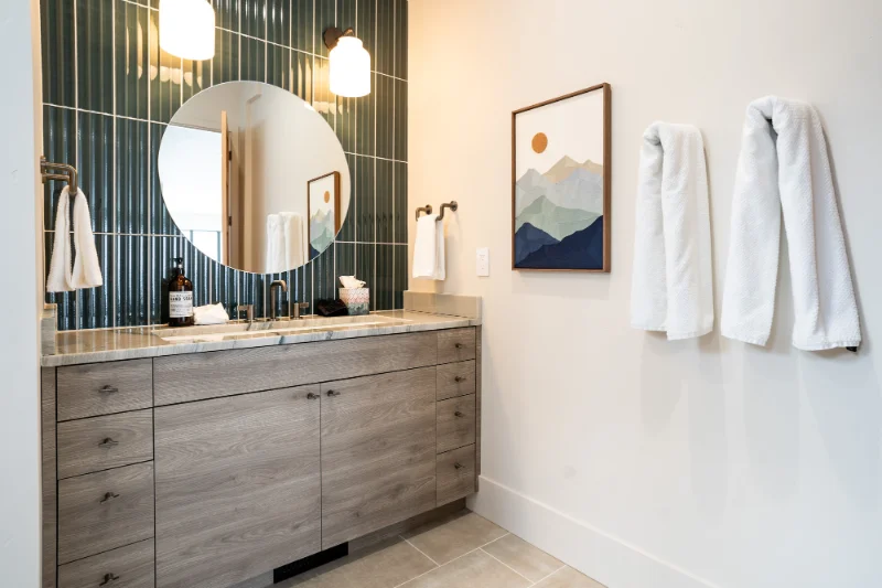 Bathroom with teal tile wall, round mirror, wood vanity, and white towels.