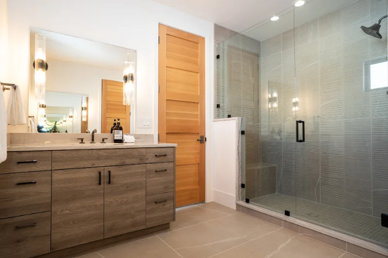 Modern bathroom with wood vanity, large mirror, glass shower, and tile floor.