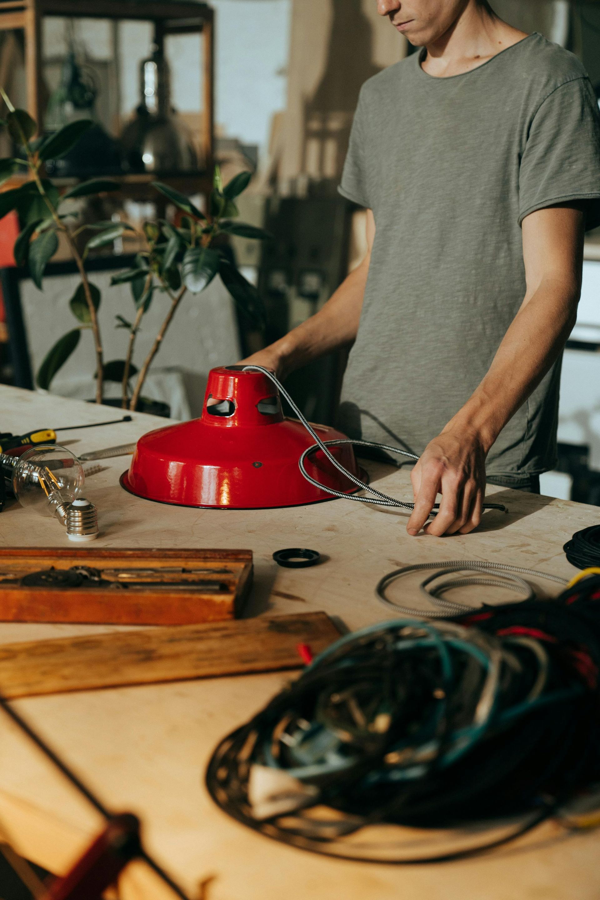 A person in a workshop assembles a bright red industrial lamp shade with a metal chain on a cluttered workbench.