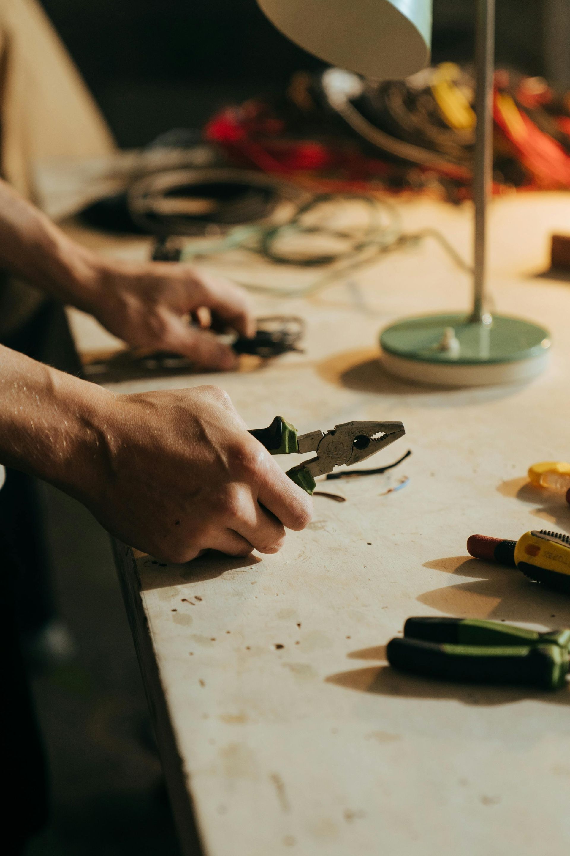 Hands holding a pair of pliers on a workbench with other tools nearby under lamp light.