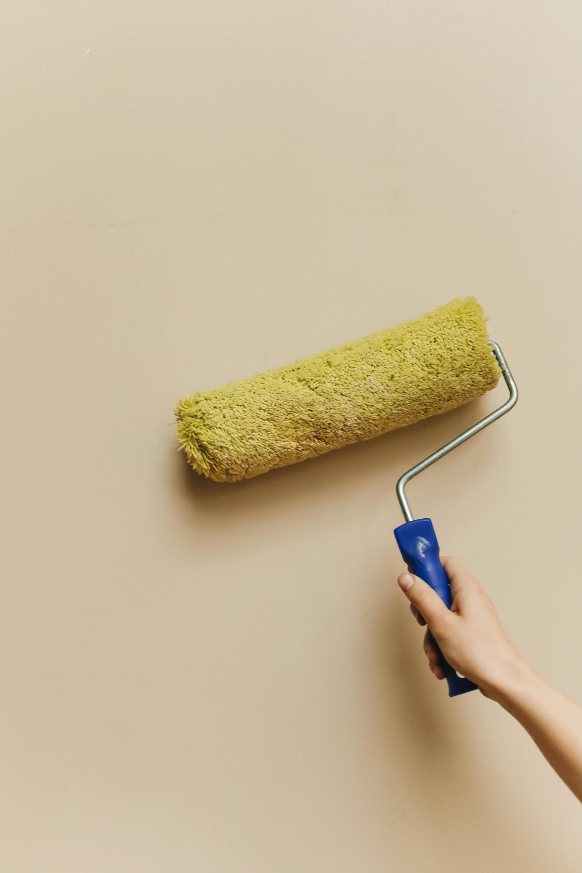 A person’s hand holds a yellow paint roller against a smooth, beige wall.