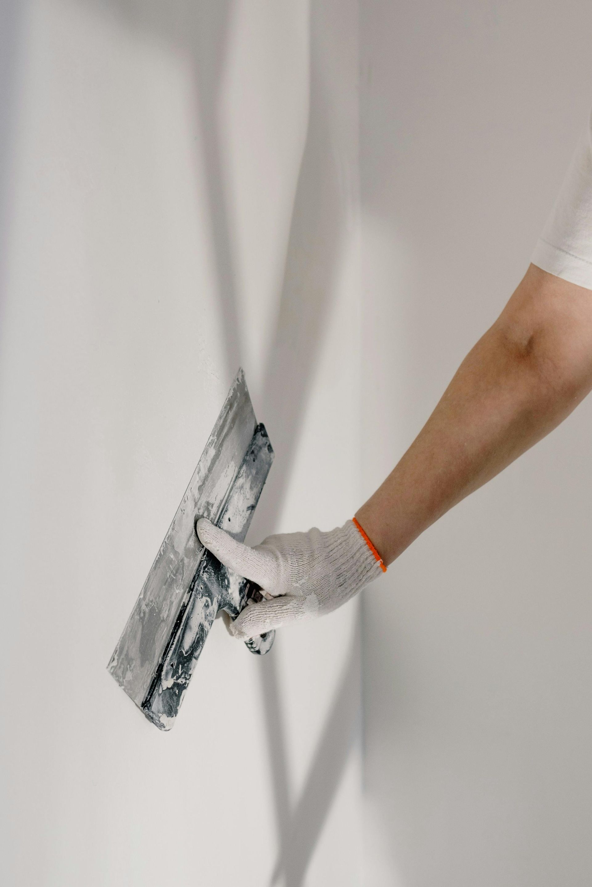 A hand in a textured work glove uses a metal putty knife to smooth white filler over a wall.