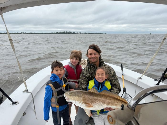 Family on a boat, posing with a large redfish caught in the water. Cloudy sky.