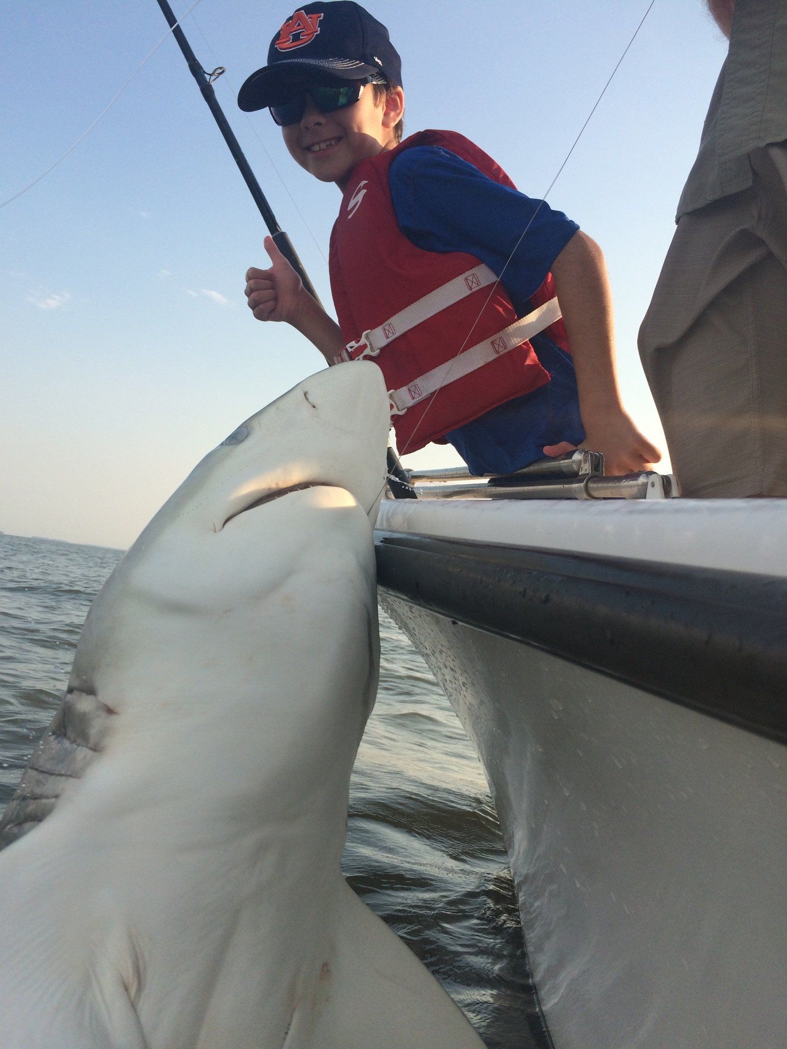 Boy on boat smiles, holding fishing rod with a shark alongside the boat; blue water.