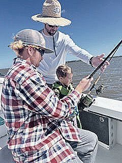 Three people fishing on a boat, one assisting a child, all wearing hats.