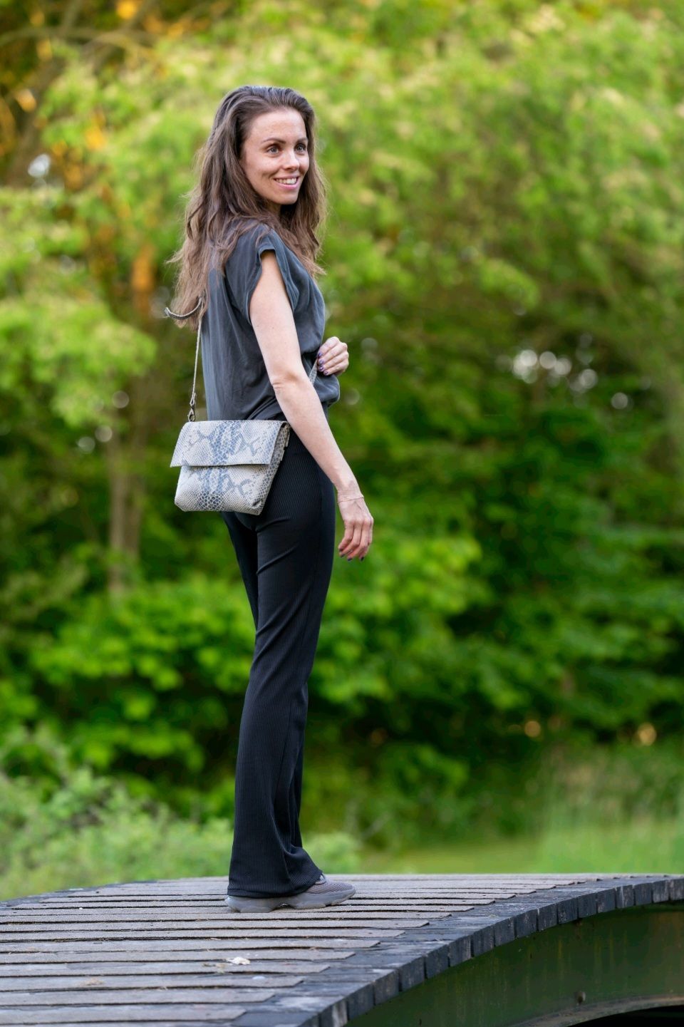 Een lachende vrouw staat op een houten brug, gekleed in een grijze top, zwarte broek en een tas met slangenprint, in de buitenlucht.