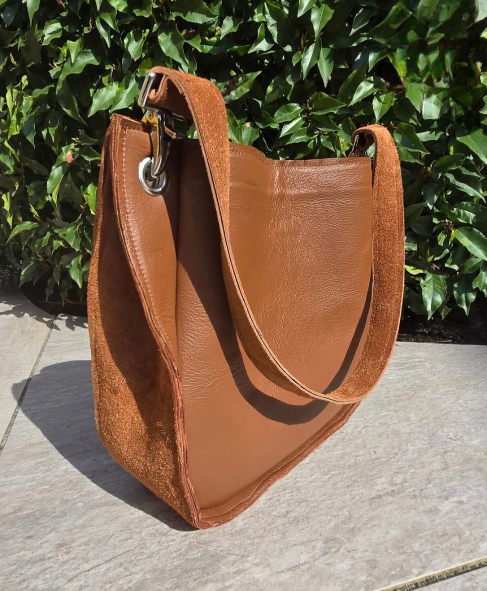 Bruine leren schoudertas met lange riem tegen een achtergrond van groene bladeren.