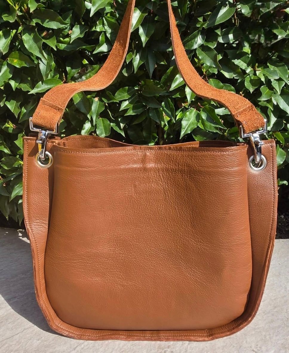 Bruine leren schoudertas met lange riem tegen een achtergrond van groene bladeren.