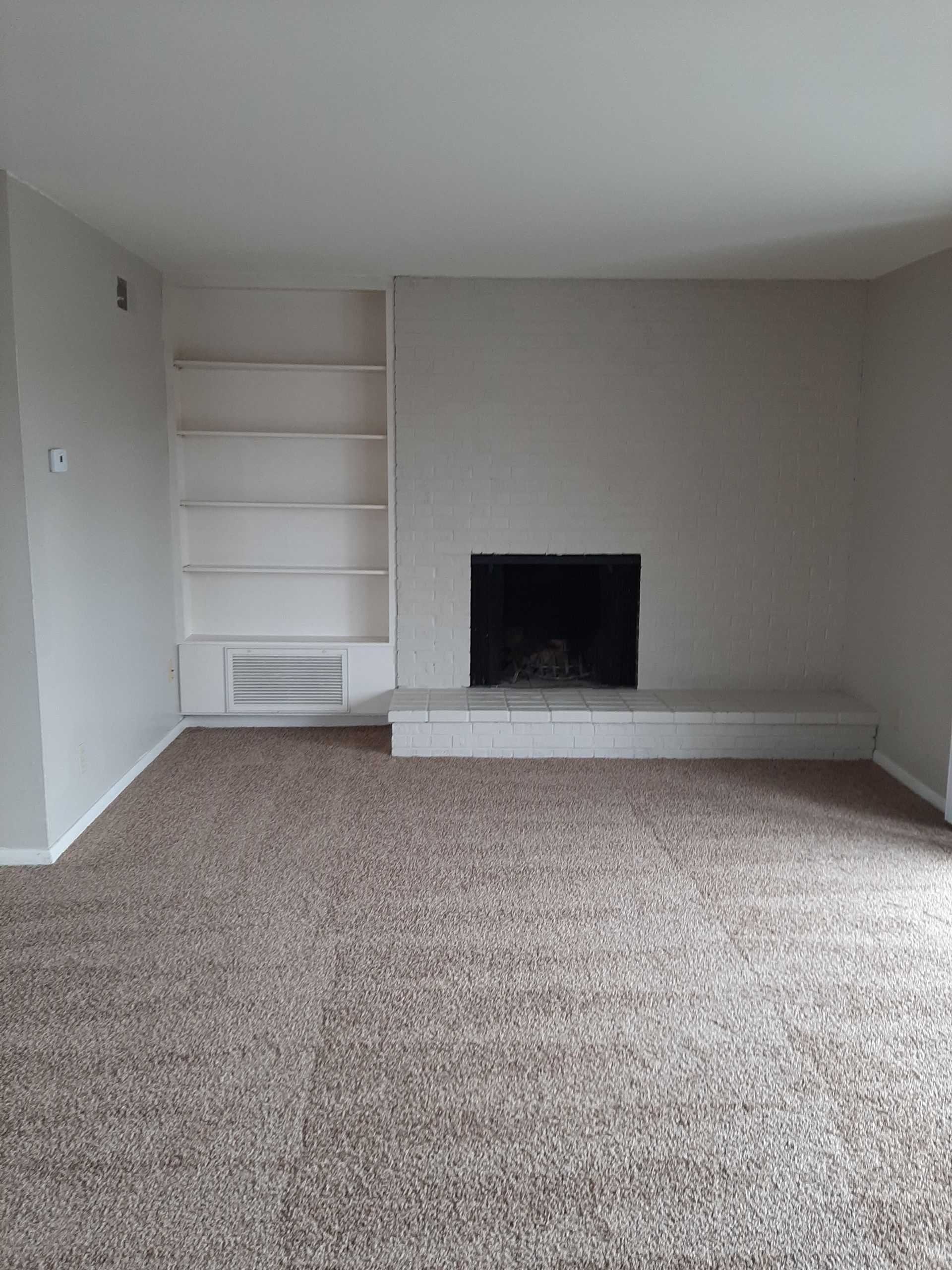 Empty living room with a fireplace, built-in shelves, and brown carpet. The walls are light grey.
