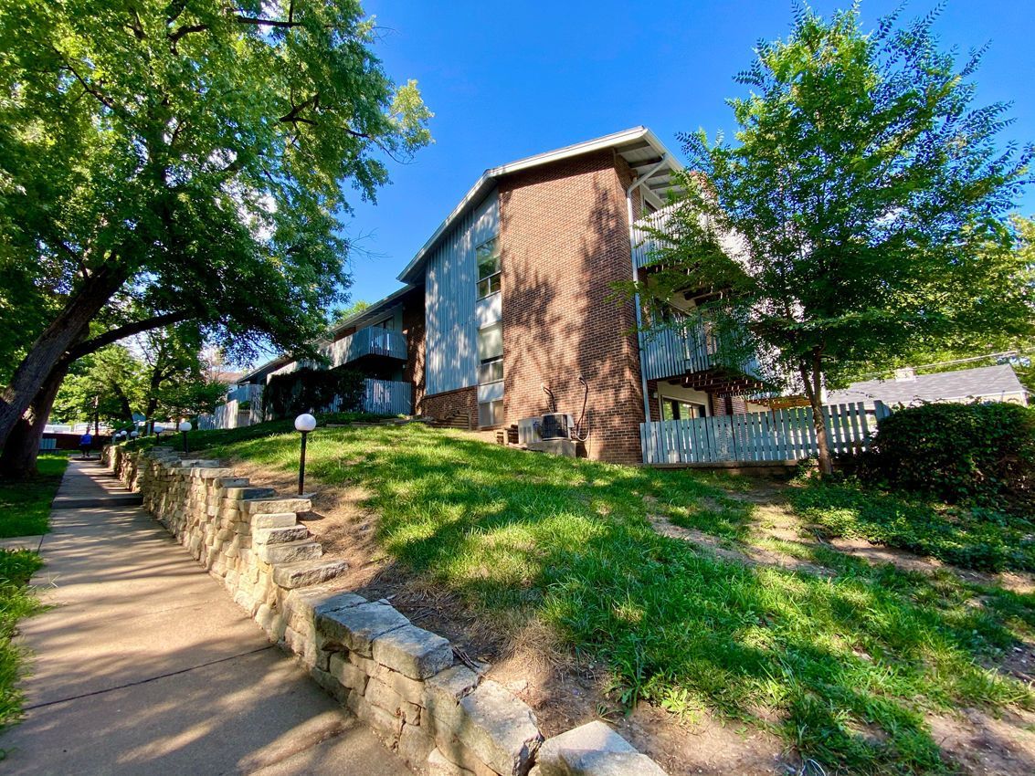 Apartment building on a slight hill with a brick facade, sidewalk, and trees under a blue sky.