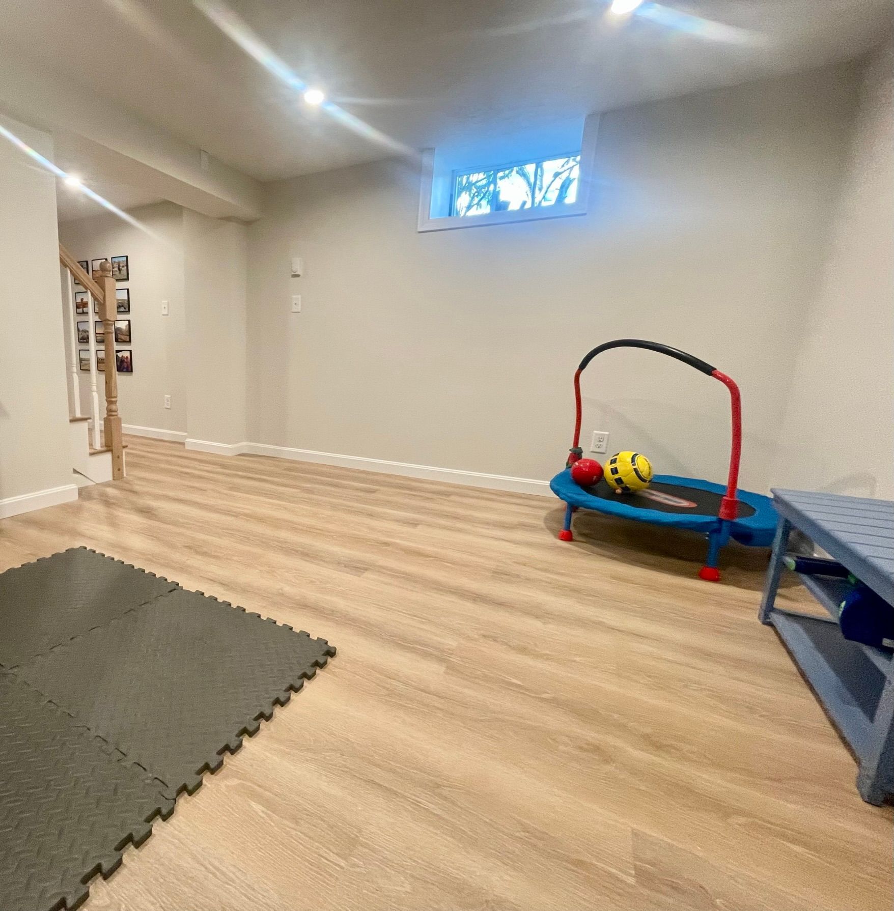 Basement play area with a trampoline, exercise mats, and a blue bench. Light wood-look flooring.