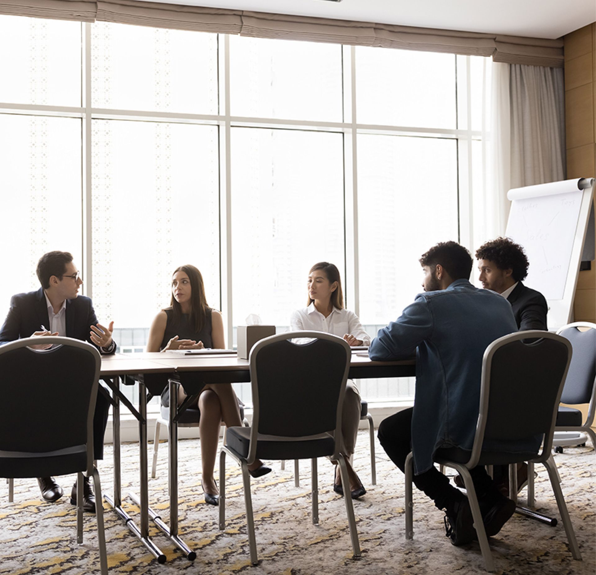Five professionals sit around a conference table in a bright, modern meeting room, engaging in a professional discussion.