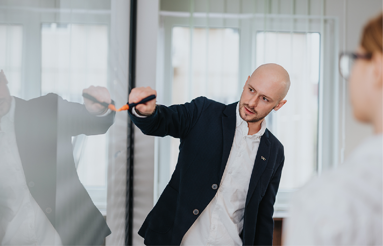 A man in a blazer writing on a glass whiteboard while facing a colleague.