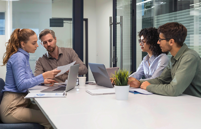 Four colleagues collaborate during a meeting around a bright conference table with laptops and documents.