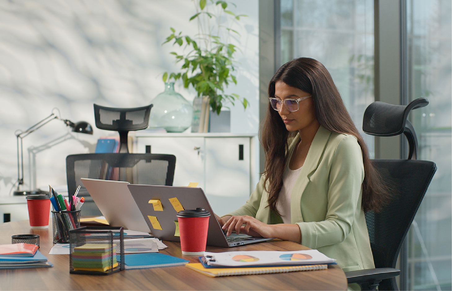 A professional woman in a light green blazer works on a laptop at a desk with a red coffee cup and office supplies.