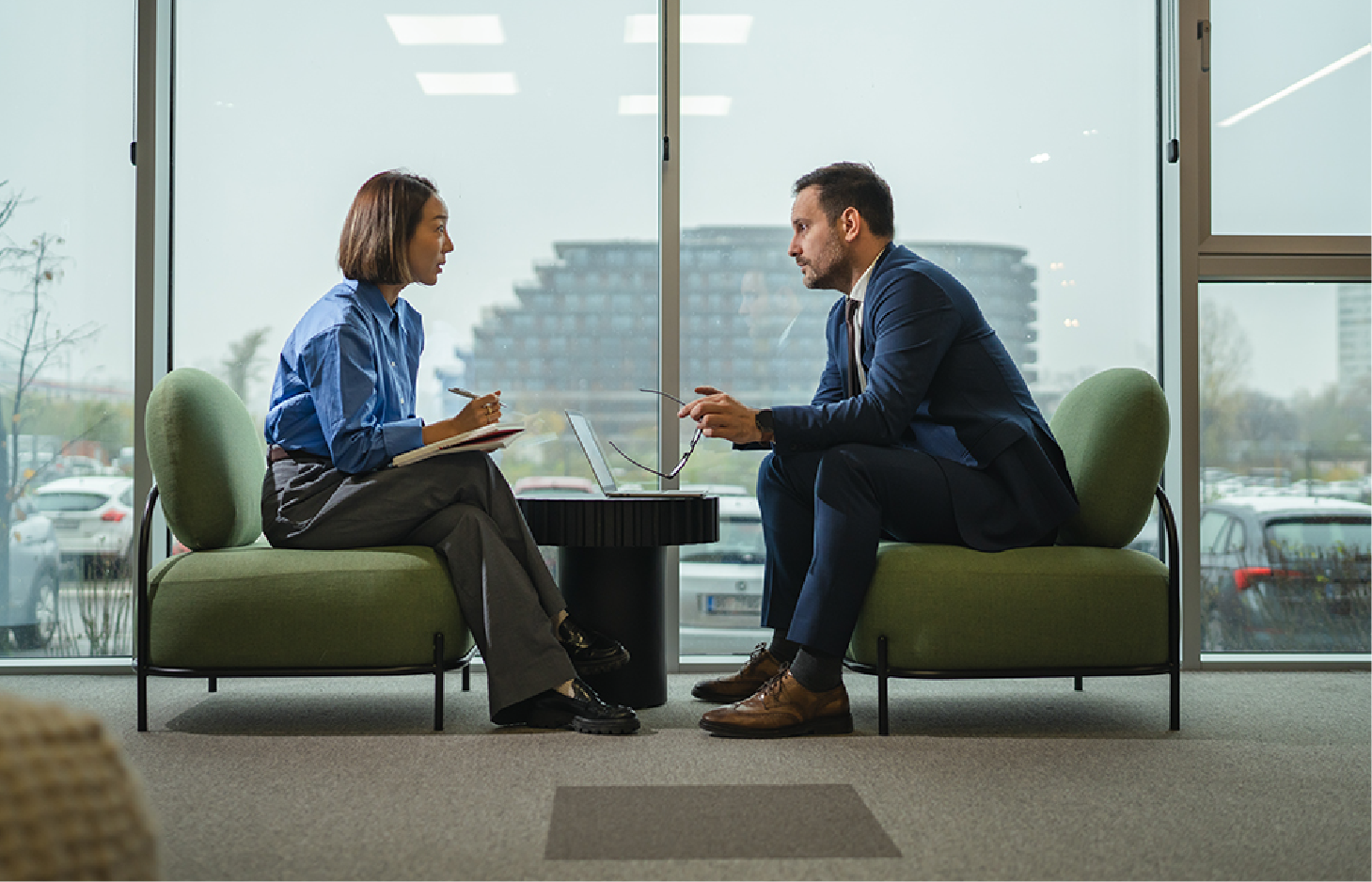 Two professionals in business attire sit in green chairs facing each other in an office with large windows.