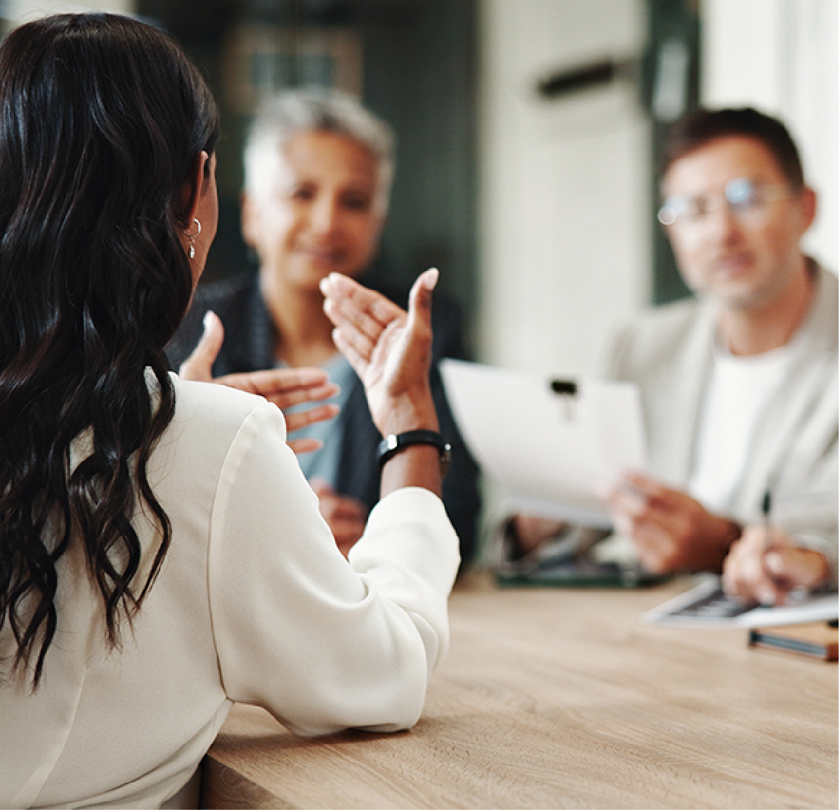 Three people in professional attire sit at a table during a meeting, with one person gesturing while speaking.