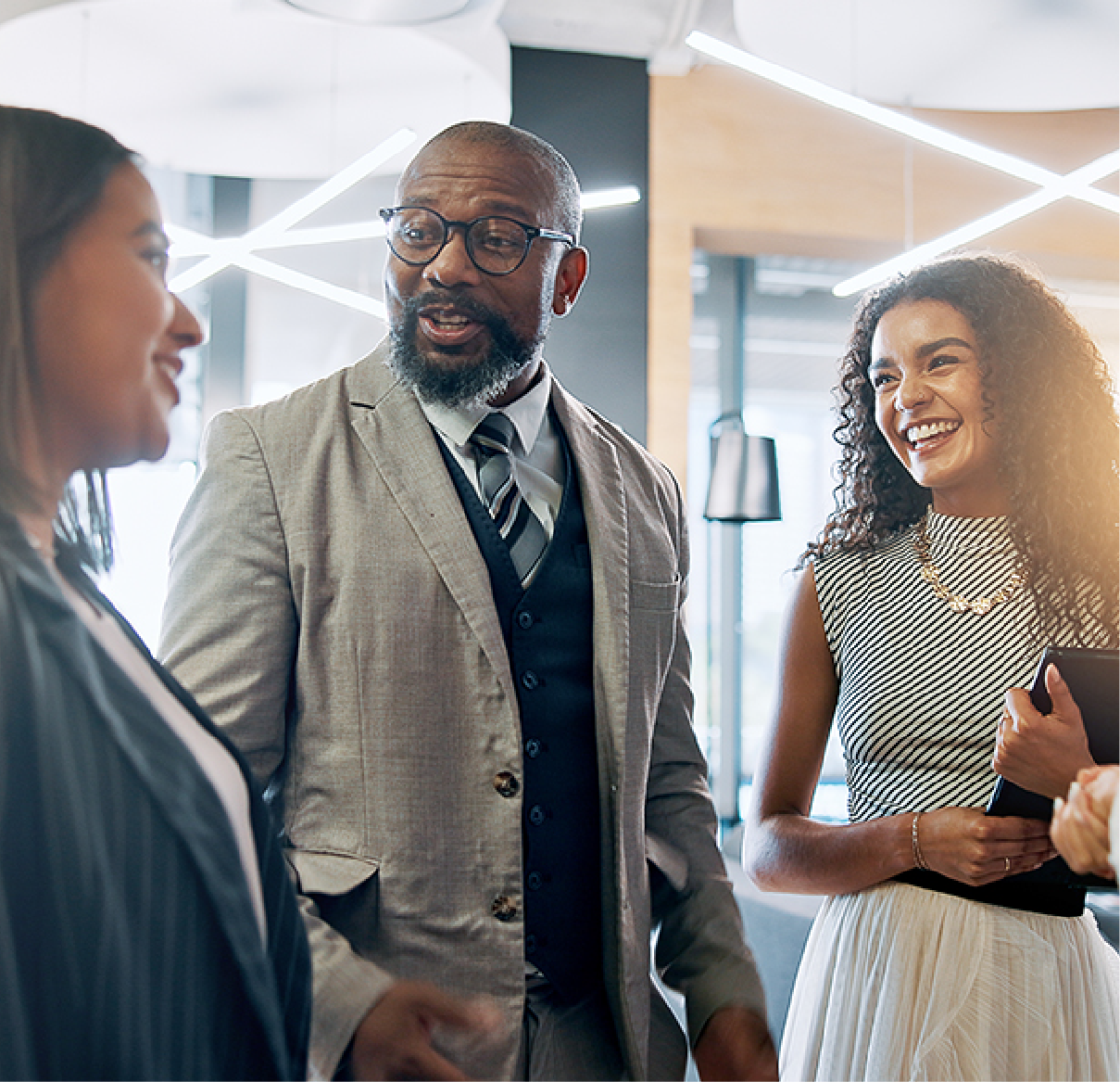 A man in a suit and vest talks with two colleagues in a brightly lit office setting, all appearing engaged and smiling.