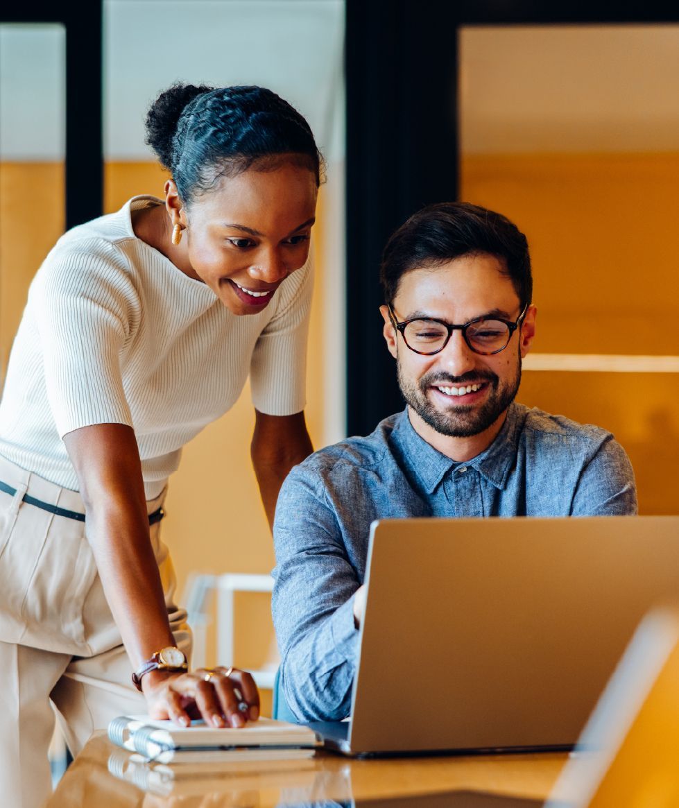 Two coworkers smile while collaborating at a laptop in a bright, modern office.