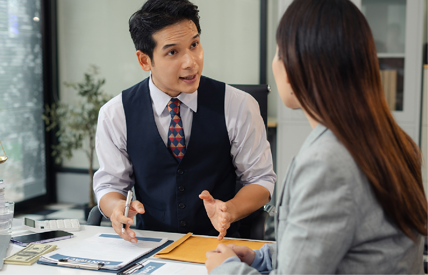 A professional in a vest and tie sits at a desk, gesturing while discussing documents with someone in an office setting.