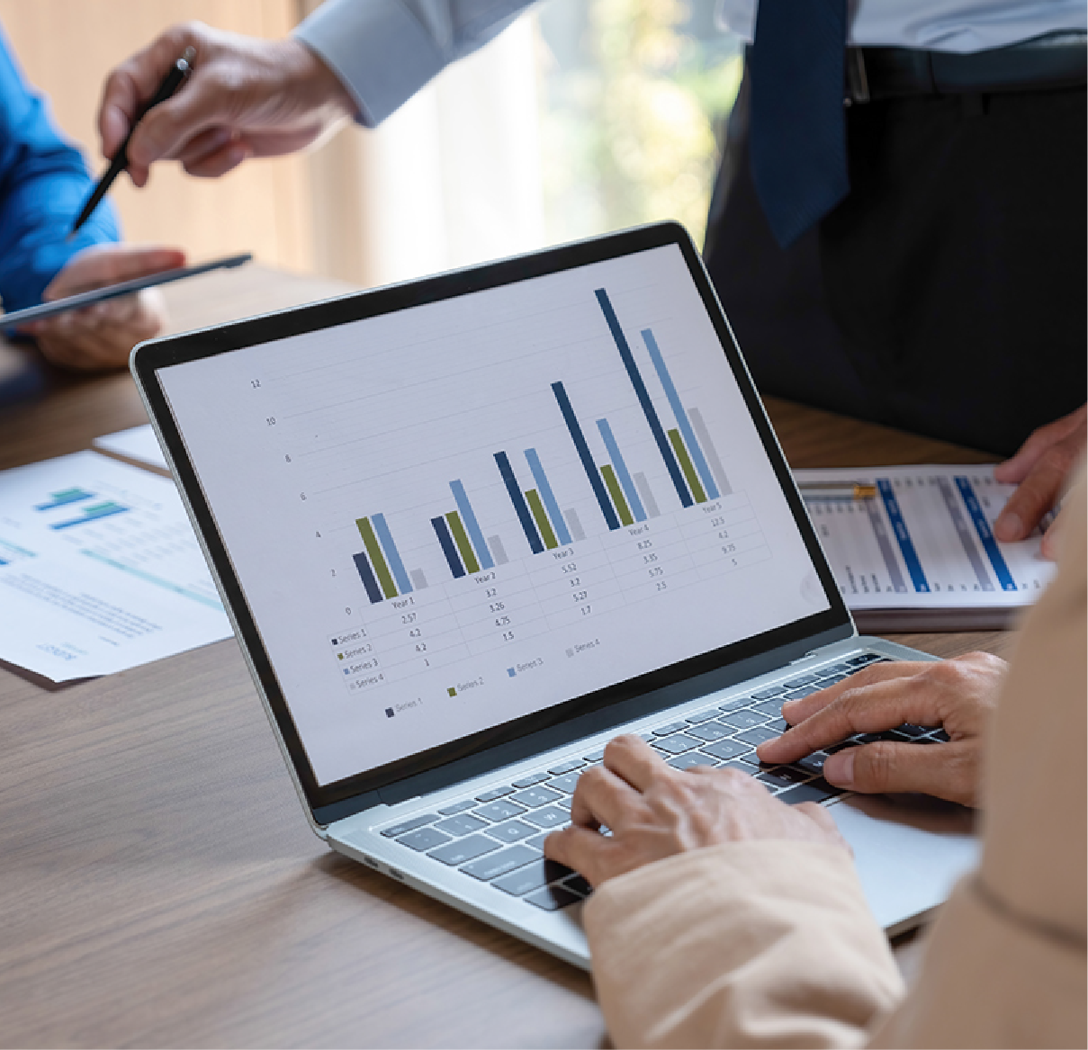 Professionals in a meeting reviewing business charts on a laptop screen and printed documents.