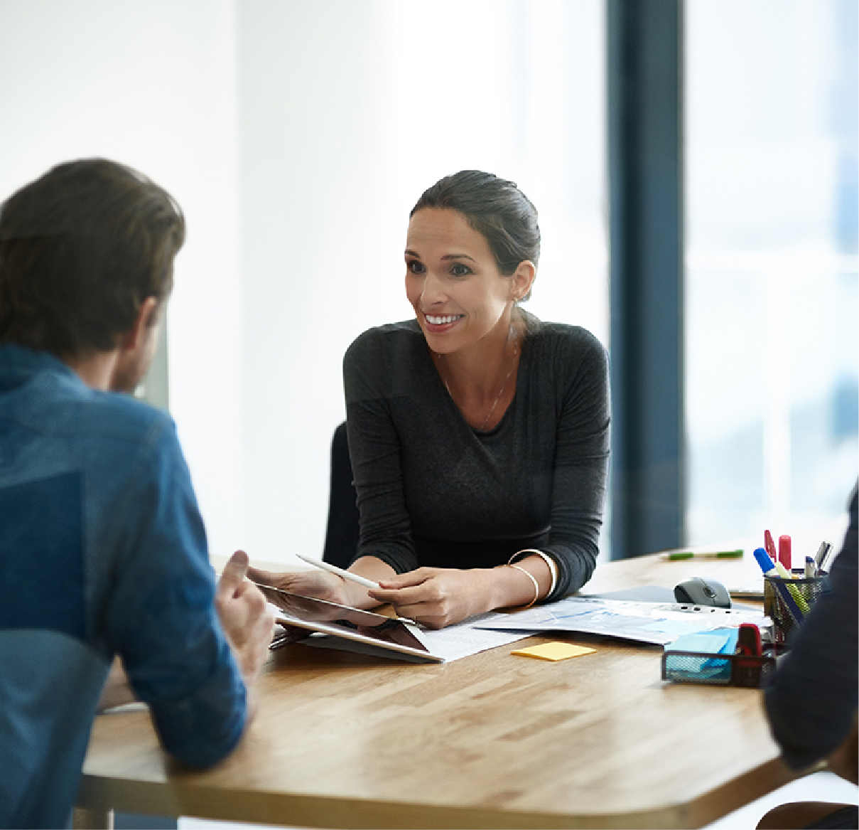 A professional woman smiles while discussing documents with a colleague across a wooden desk in a bright office.