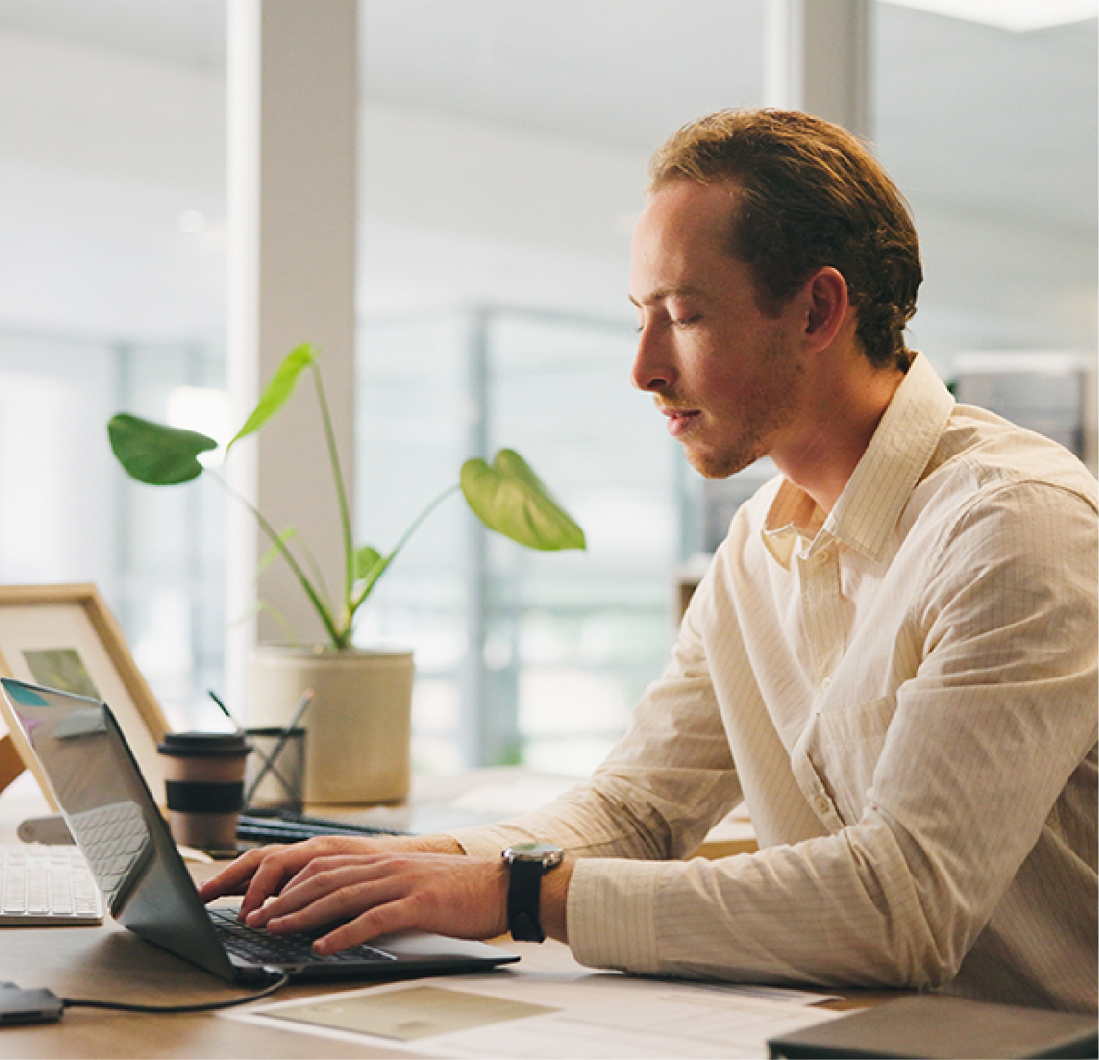 A person in a light-colored shirt typing on a laptop at a bright office desk with a potted plant.