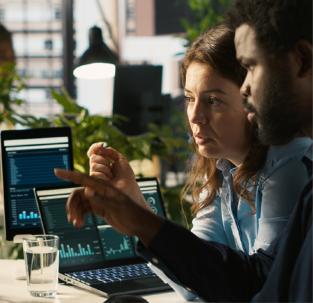 Two colleagues collaborate in a brightly lit office, analyzing business data displayed on laptop and tablet screens.