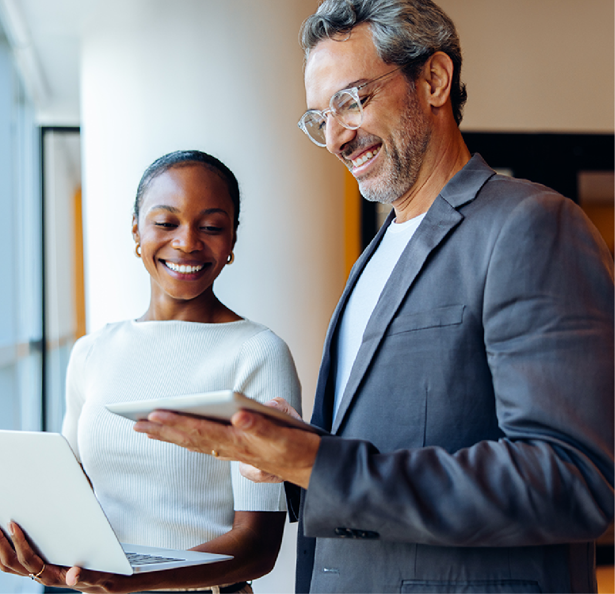 Two professionals stand side-by-side, one holding a laptop and the other a tablet, both smiling while collaborating.
